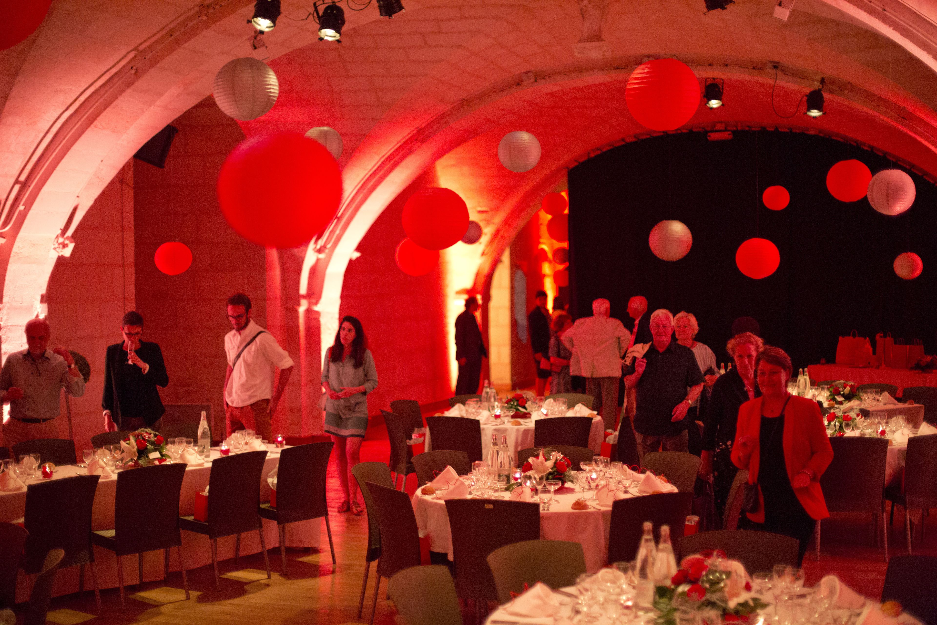 A banquet hall with round tables set for dining, a stage in the background, and red lighting.