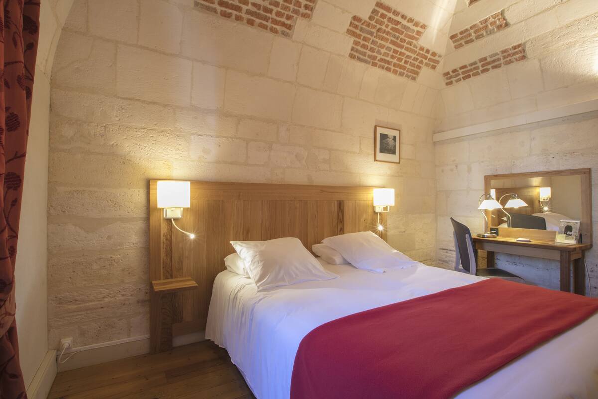 Une chambre d’hôtel avec un plafond orné d’un motif en briques, une tête de lit en bois, un lit avec une literie blanche, un plaid rouge, un bureau avec une lampe et un miroir.
