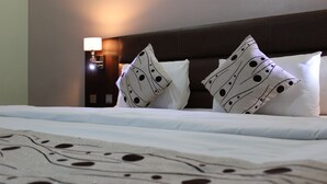Habitación de hotel con una cama grande, dos almohadones decorativos, una mesita de noche con lámpara y un reloj de pared.