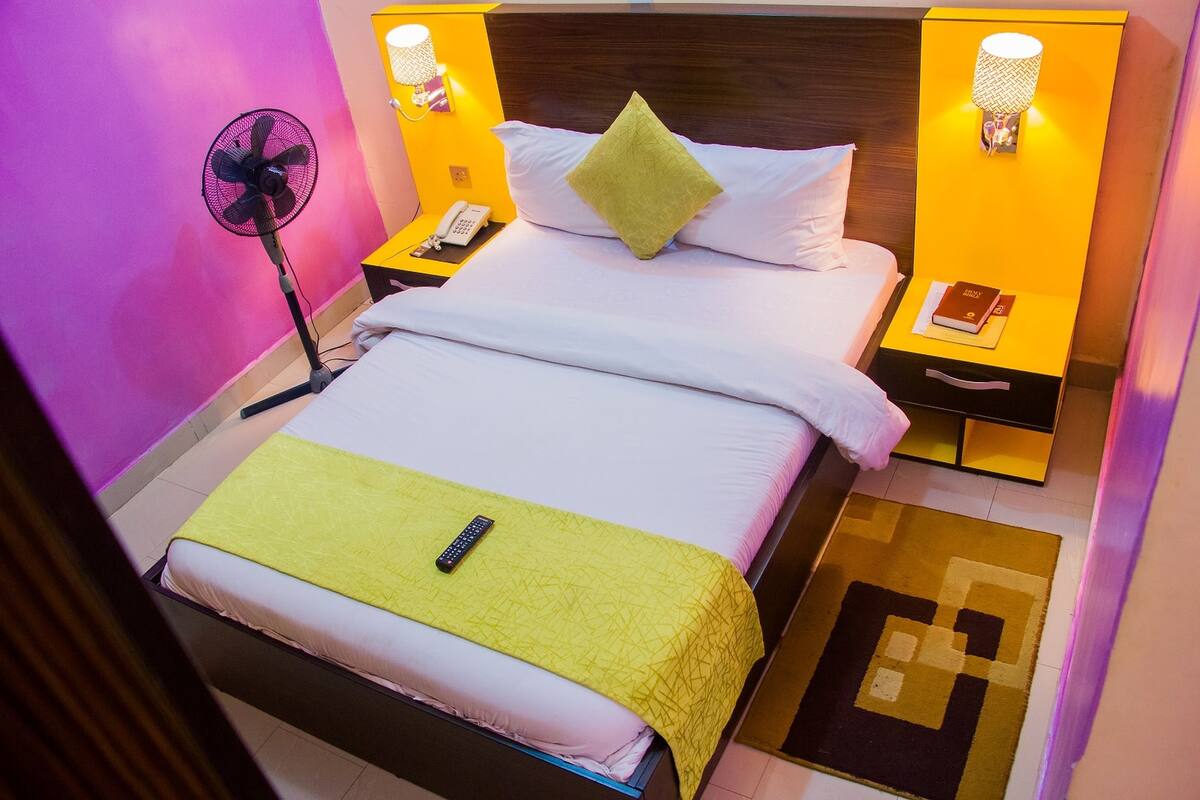 A hotel room with a bed, a bedside table with a lamp, a fan, and a phone.
