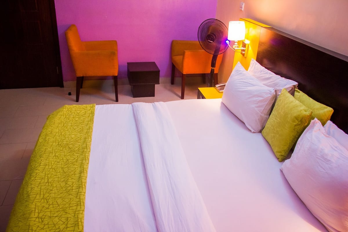 A hotel room with a bed, two chairs, a small table, and a wall-mounted fan.