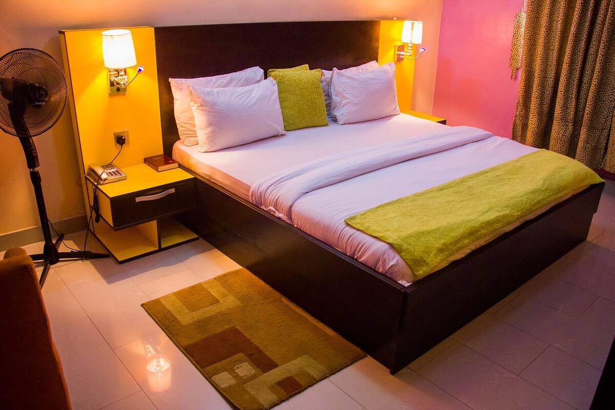 A hotel room with a large bed, bedside lamps, a nightstand, a fan, and a rug.