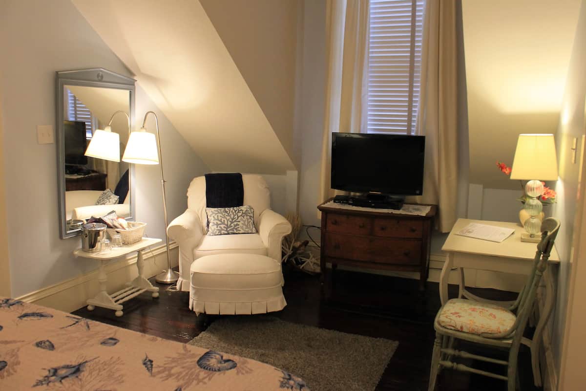 A bedroom with a bed, a chair, a desk, a television, a mirror, and a window with blinds.