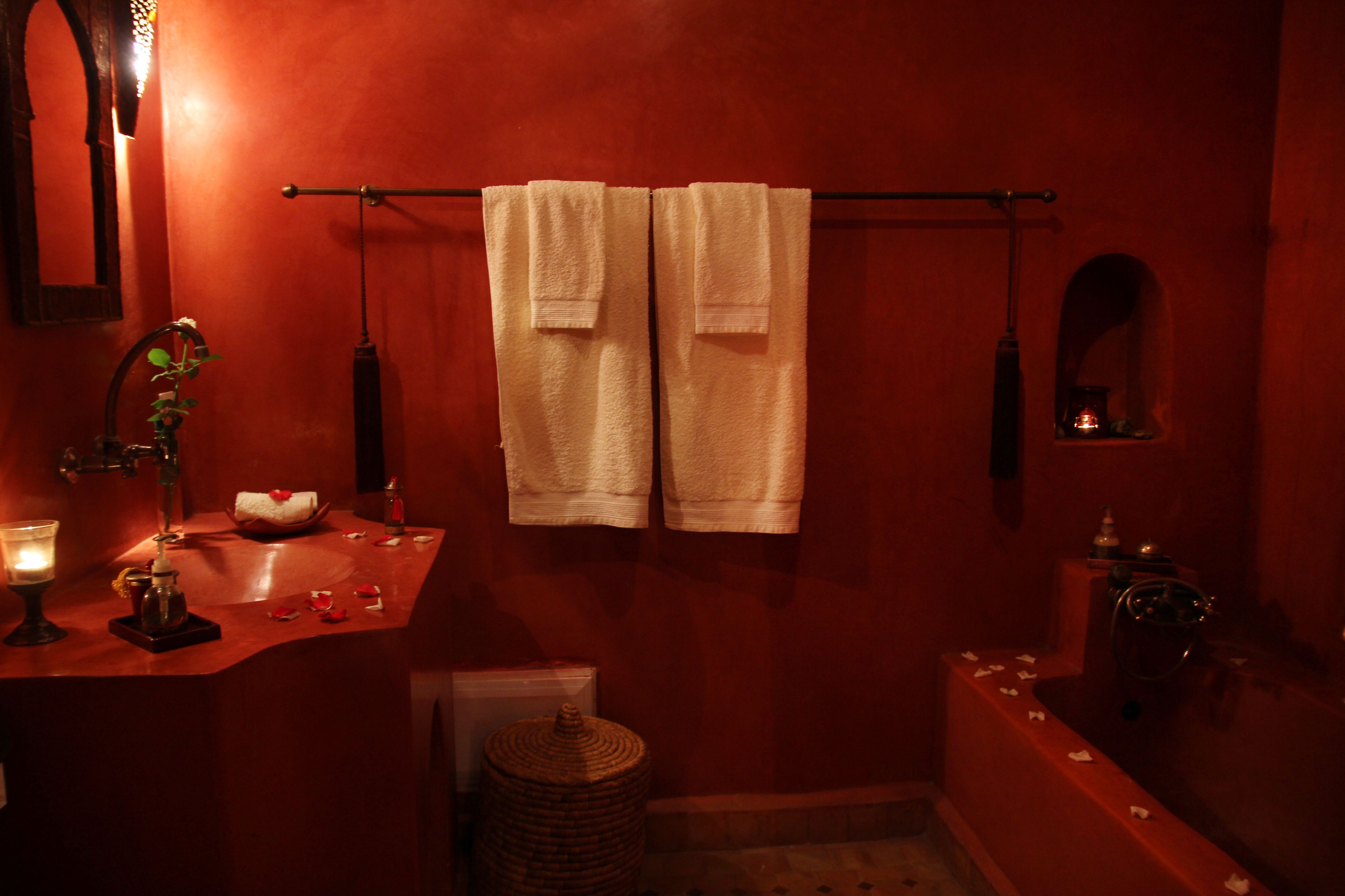 A bathroom with a red wall, a sink, a mirror, a towel rack with two towels, a candle, and a small window.
