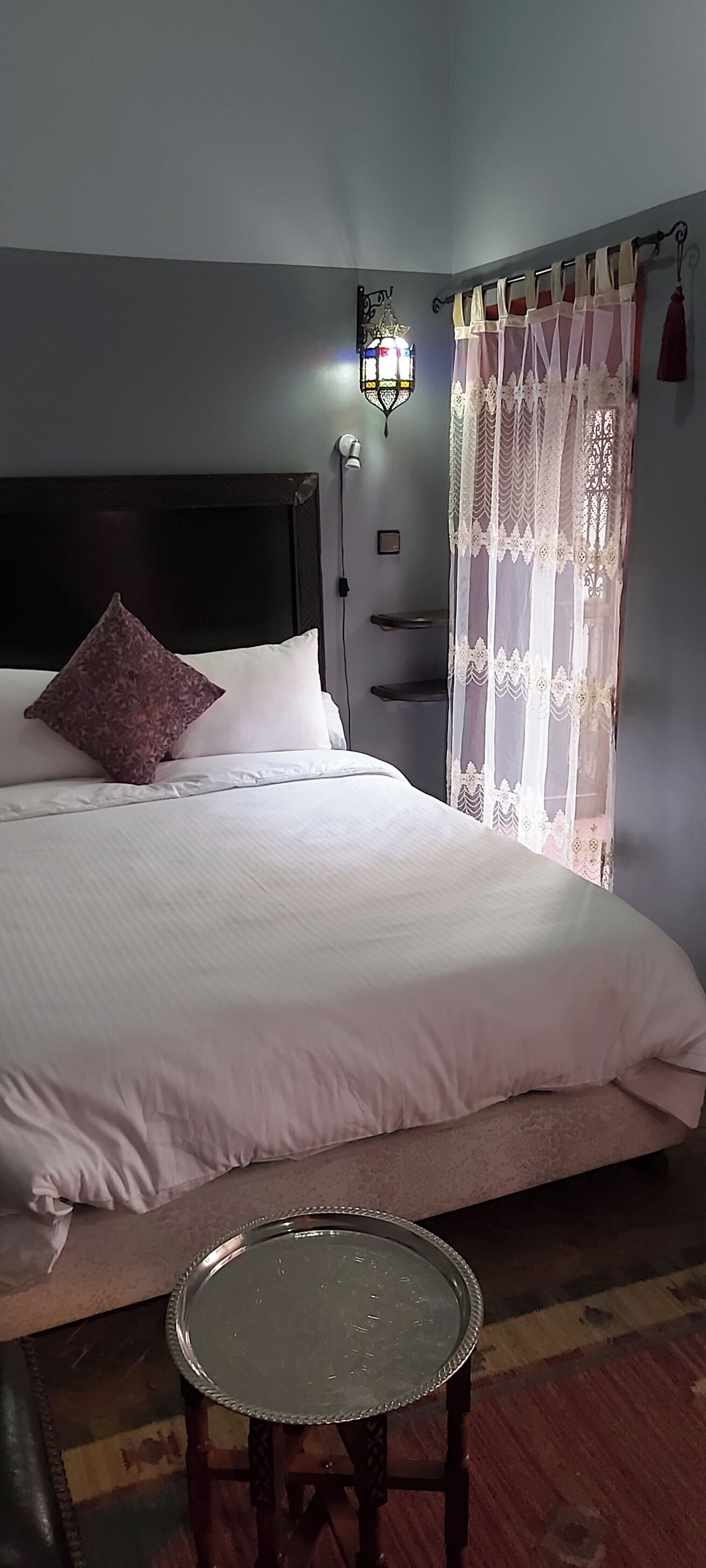 A neatly made bed with a headboard, a bedside table, and a curtain with a pattern.