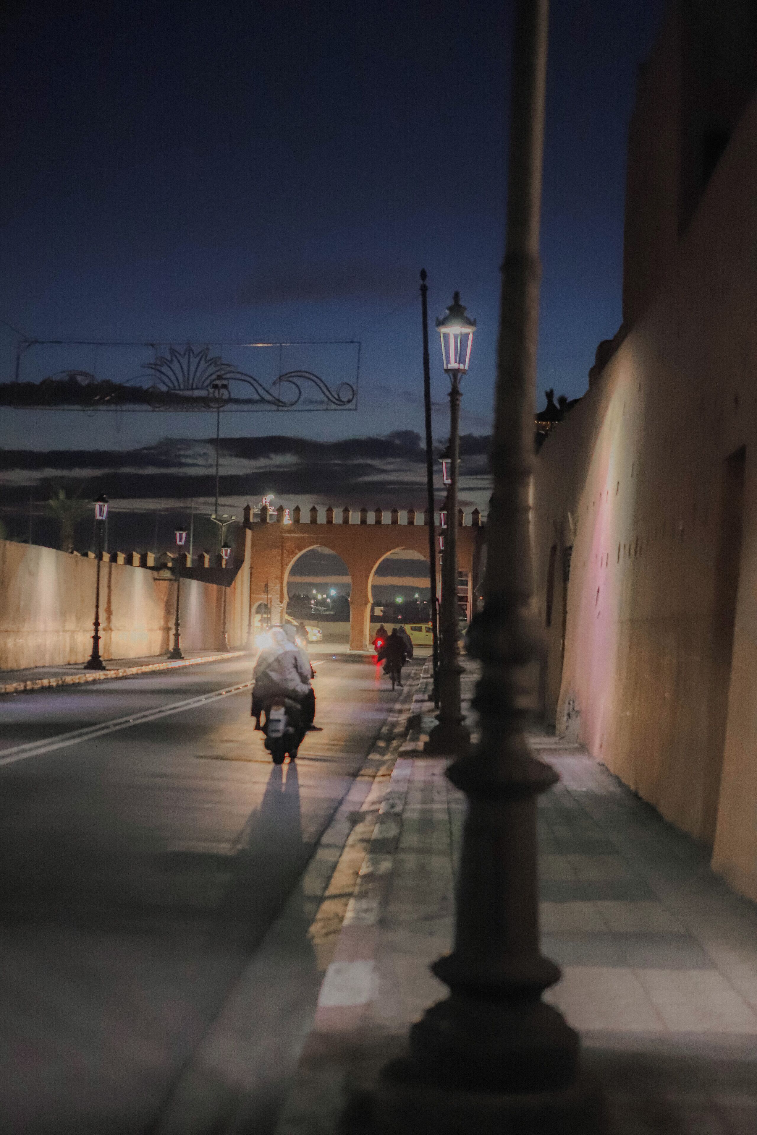 Ein belebter Straßenansicht bei Nacht mit einer Fußgängerbrücke, Straßenlaternen und einem Motorradfahrer.