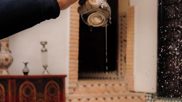 A person pouring tea from a traditional glass jug onto a plate with a cup. - Riad Ajebel (Marrakech)