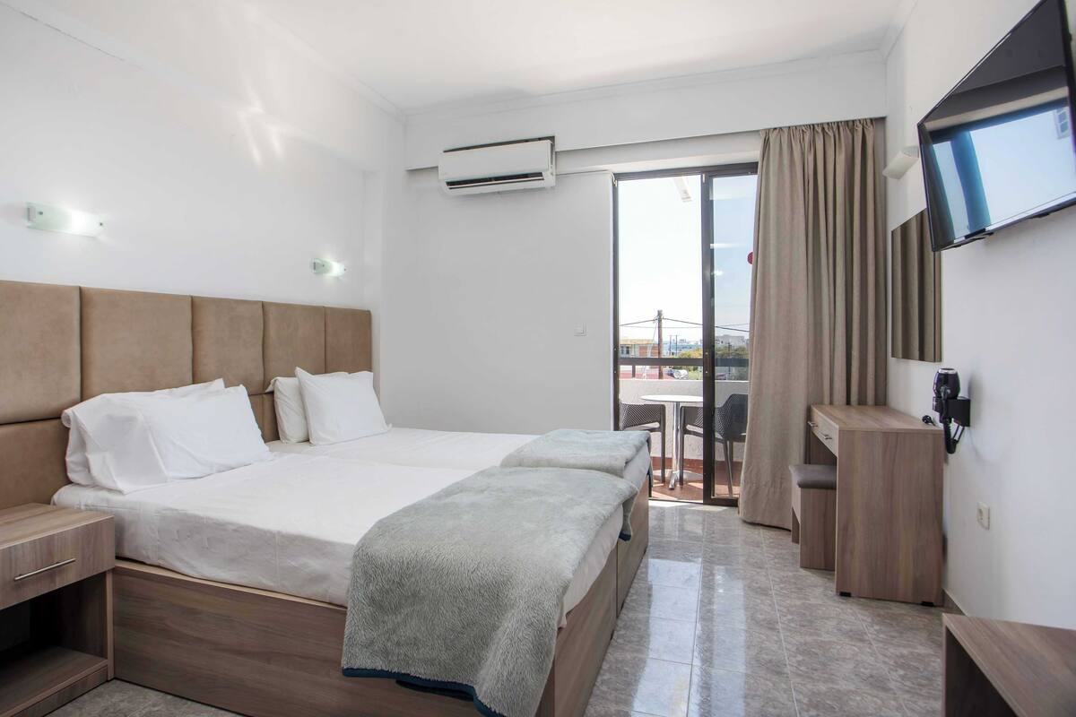 A hotel room with a double bed, a desk, a TV, an air conditioner, and a view of the outside.