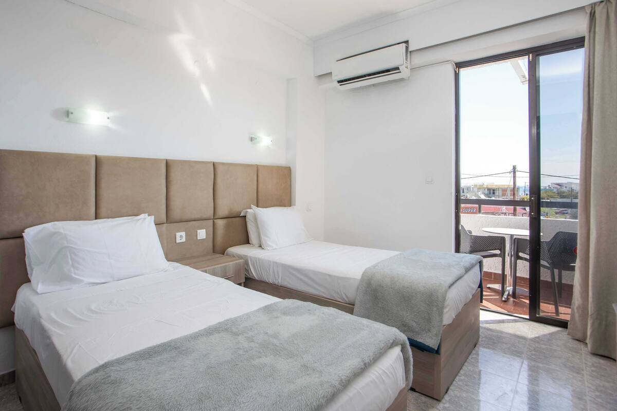 A hotel room with two beds, a balcony with a table and chairs, and an air conditioning unit.