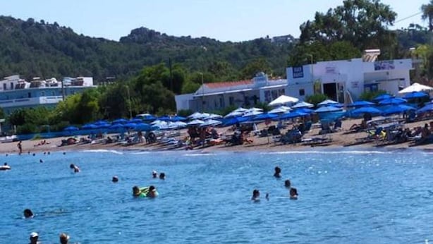 Uma praia com pessoas nadando e tomando sol, uma fileira de guarda-sóis azuis e edifícios ao fundo.