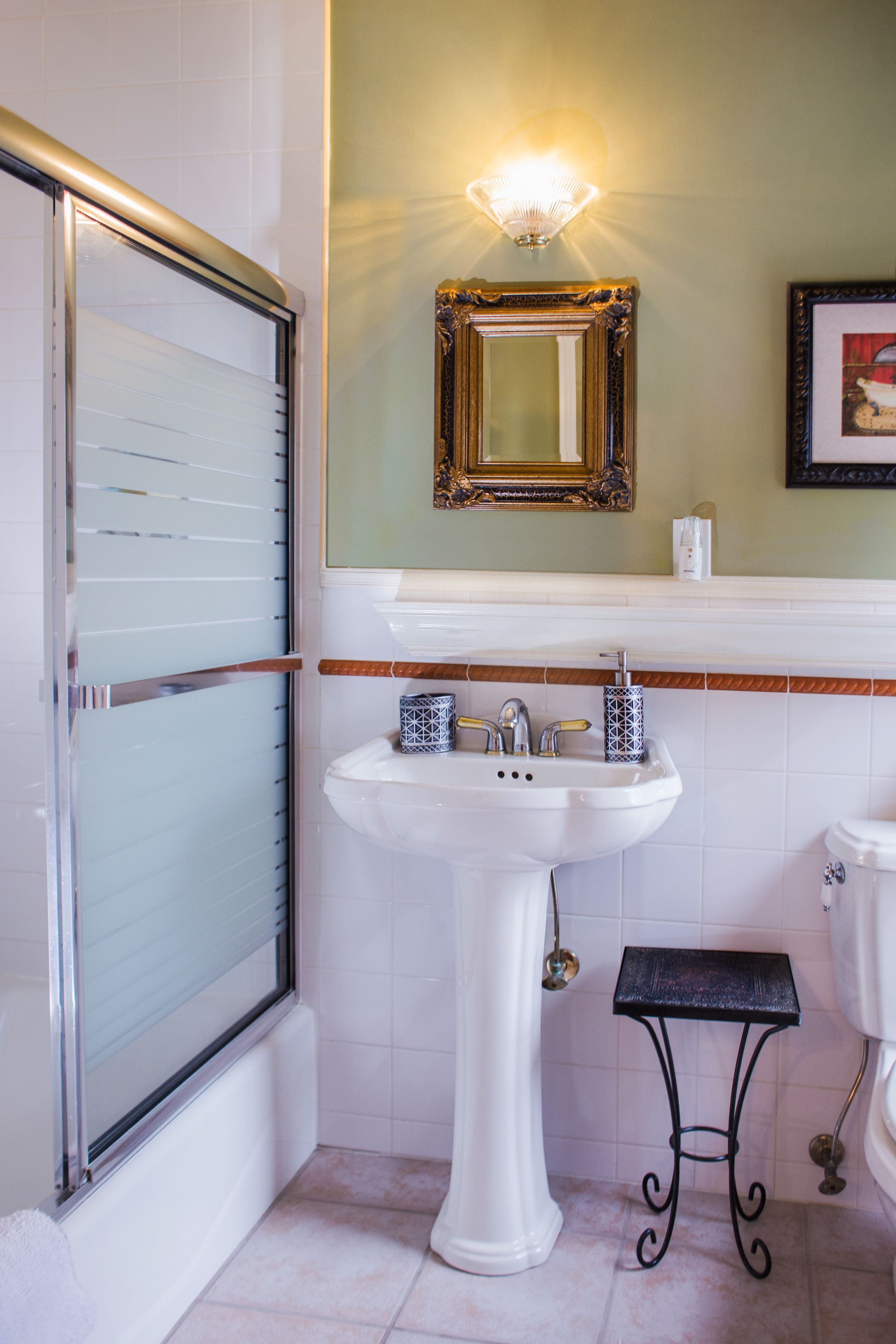 A bathroom with a glass shower enclosure, a pedestal sink, a small black stool, and a framed mirror on the wall.