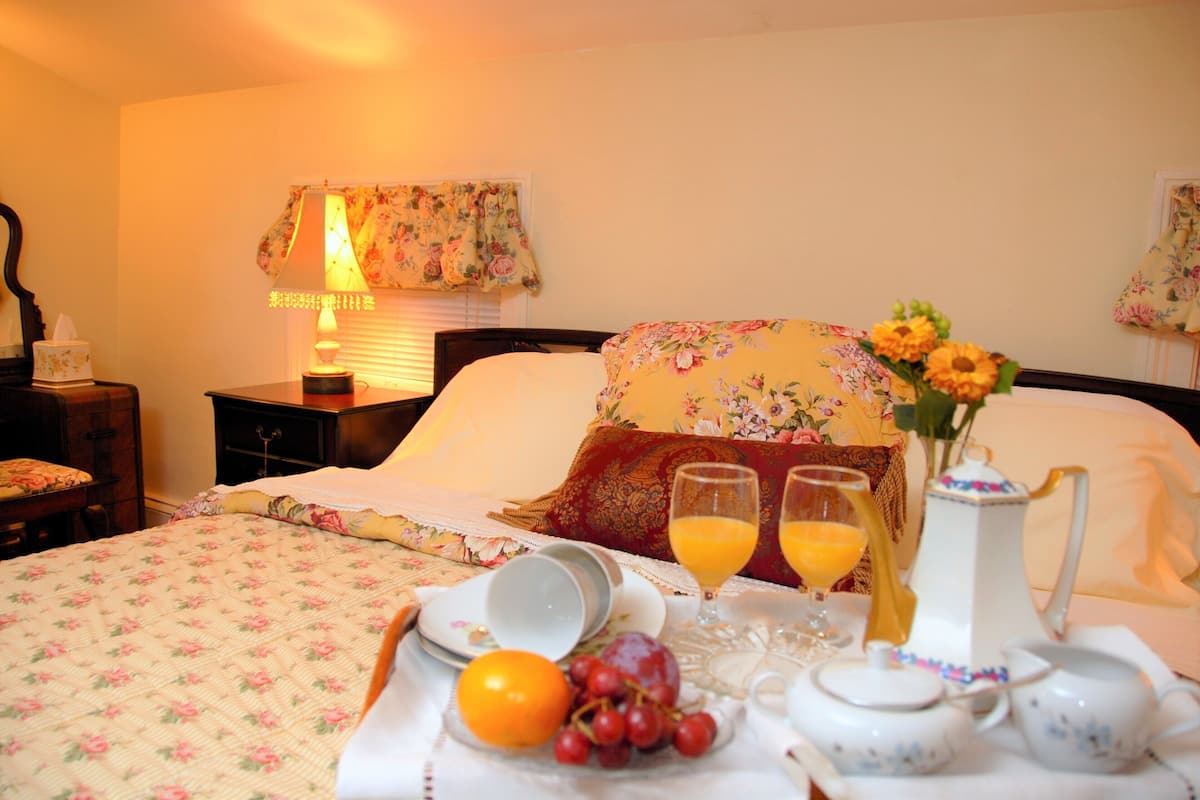 A neatly made bed with floral bedding, a bedside table with a lamp, and a breakfast tray with fruits and a pitcher.