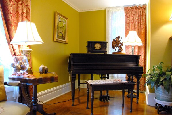 A room with a piano, a table with a lamp, a chair, and a potted plant.