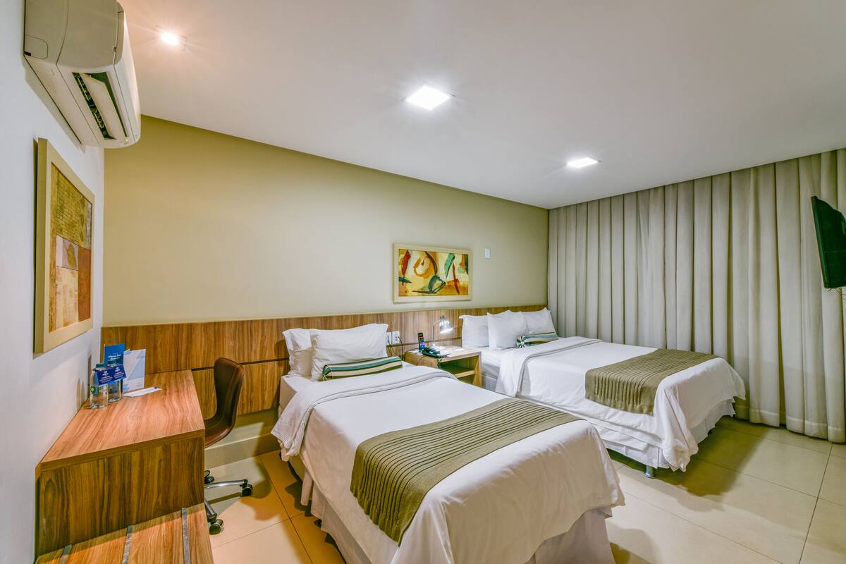 A hotel room with two beds, a desk, a chair, an air conditioner, and a painting on the wall.