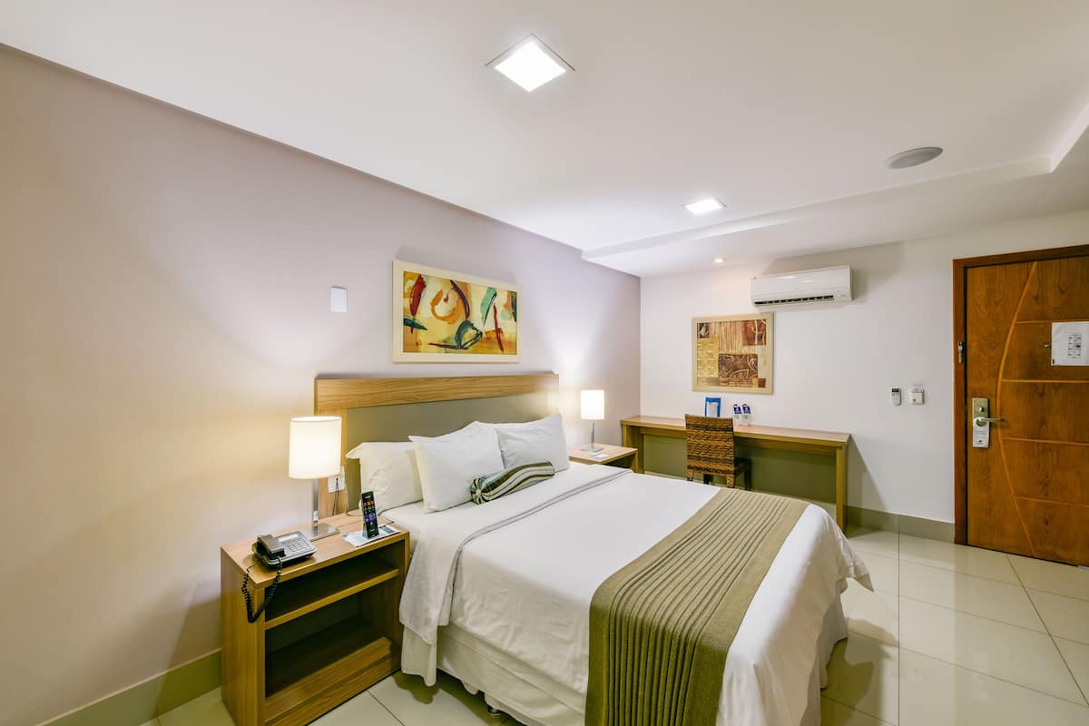 A hotel room with a bed, bedside tables, a lamp, an air conditioning unit, and artwork on the walls.