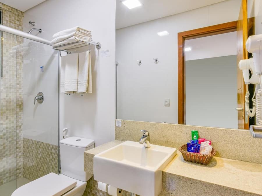 Un baño con inodoro, lavabo y ducha. Hay un perchero con toallas, un bote de basura y una cesta de artículos de aseo.