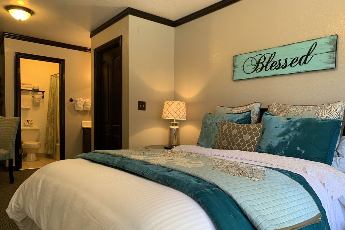 Ein ordentlich bezogenes Bett mit einem türkisfarbenen und weißen Bettüberwurf, ein Schild an der Wand mit der Aufschrift "Blessed" und ein durch eine offene Tür einsehbarer Badezimmer.