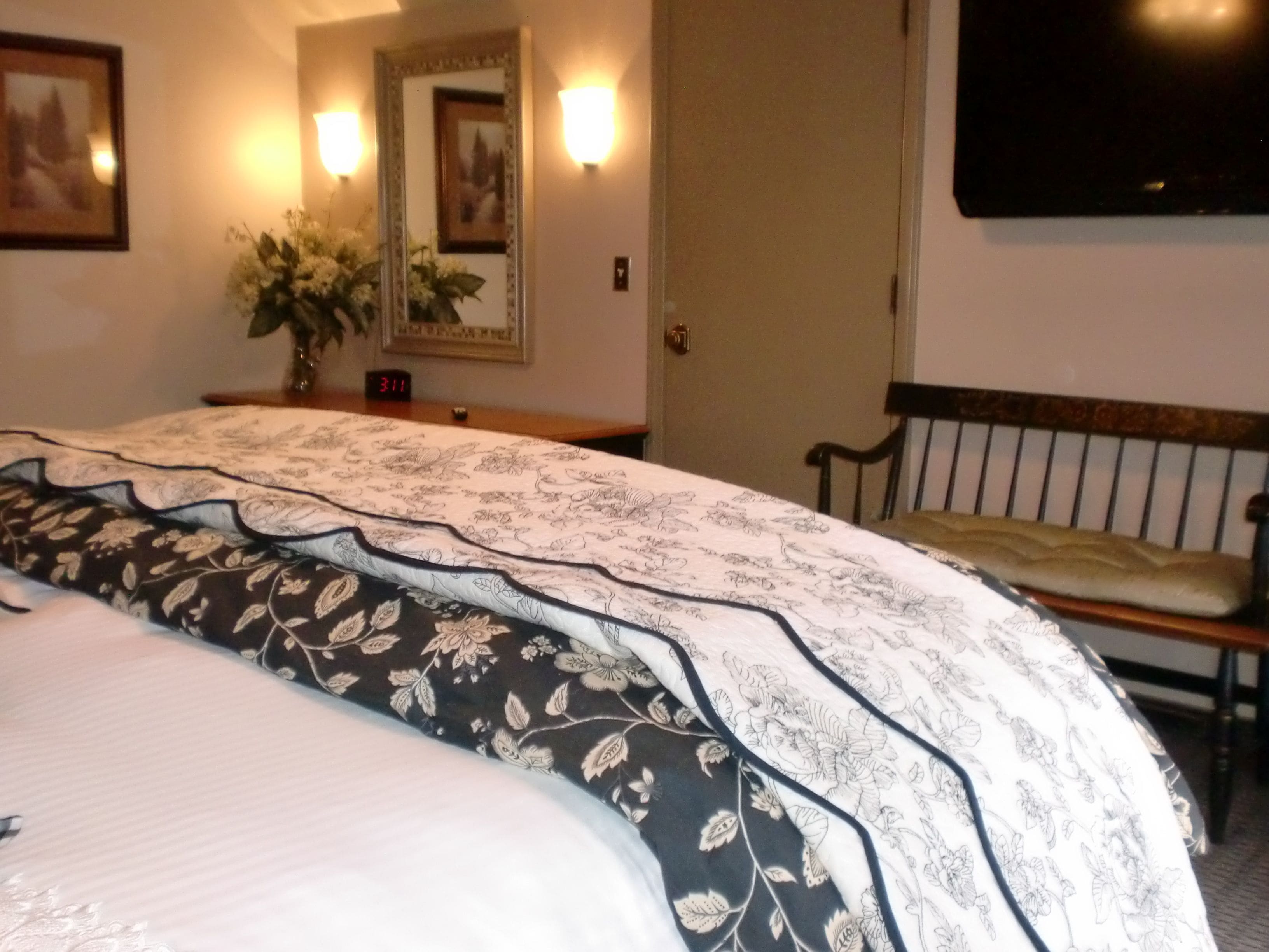 Una habitación de hotel con una cama grande, una mesita de noche con un reloj, un espejo, un televisor montado en la pared y un banco.
