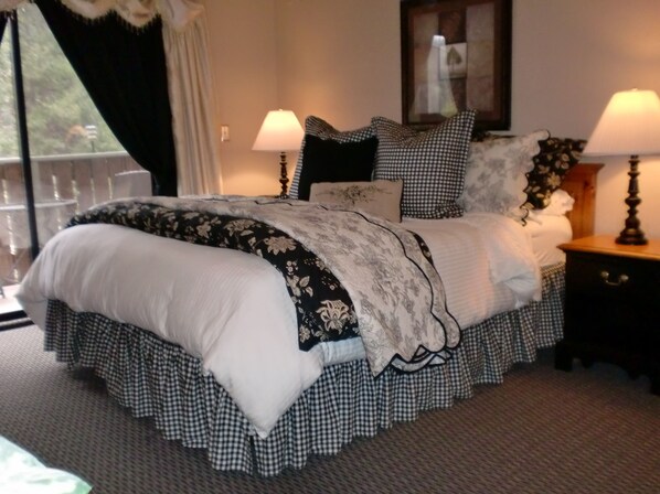 A neatly made bed with a floral-patterned bedspread and checkered pillows, two bedside lamps, and a nightstand with a lamp. - The North Shore Inn (Crestline)