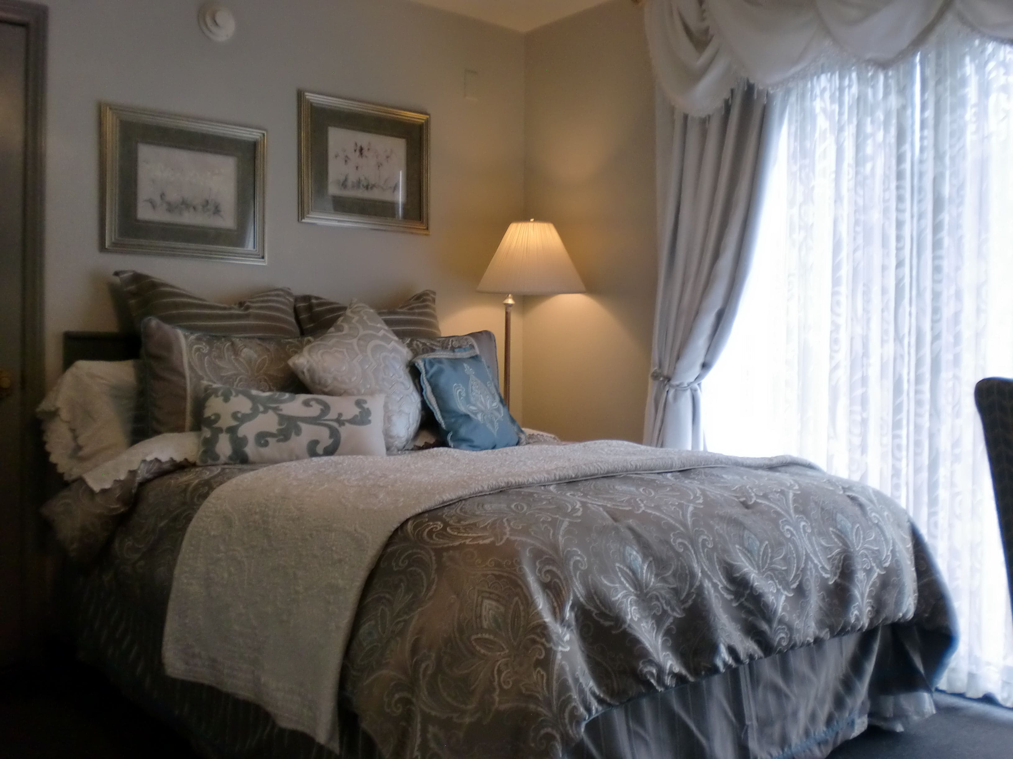 Una cama bien hecha con una colcha estampada y varias almohadas. Dos cuadros enmarcados decoran la pared sobre la cama. Una lámpara está en el suelo junto a la cama, y se ve una ventana con cortinas al fondo.