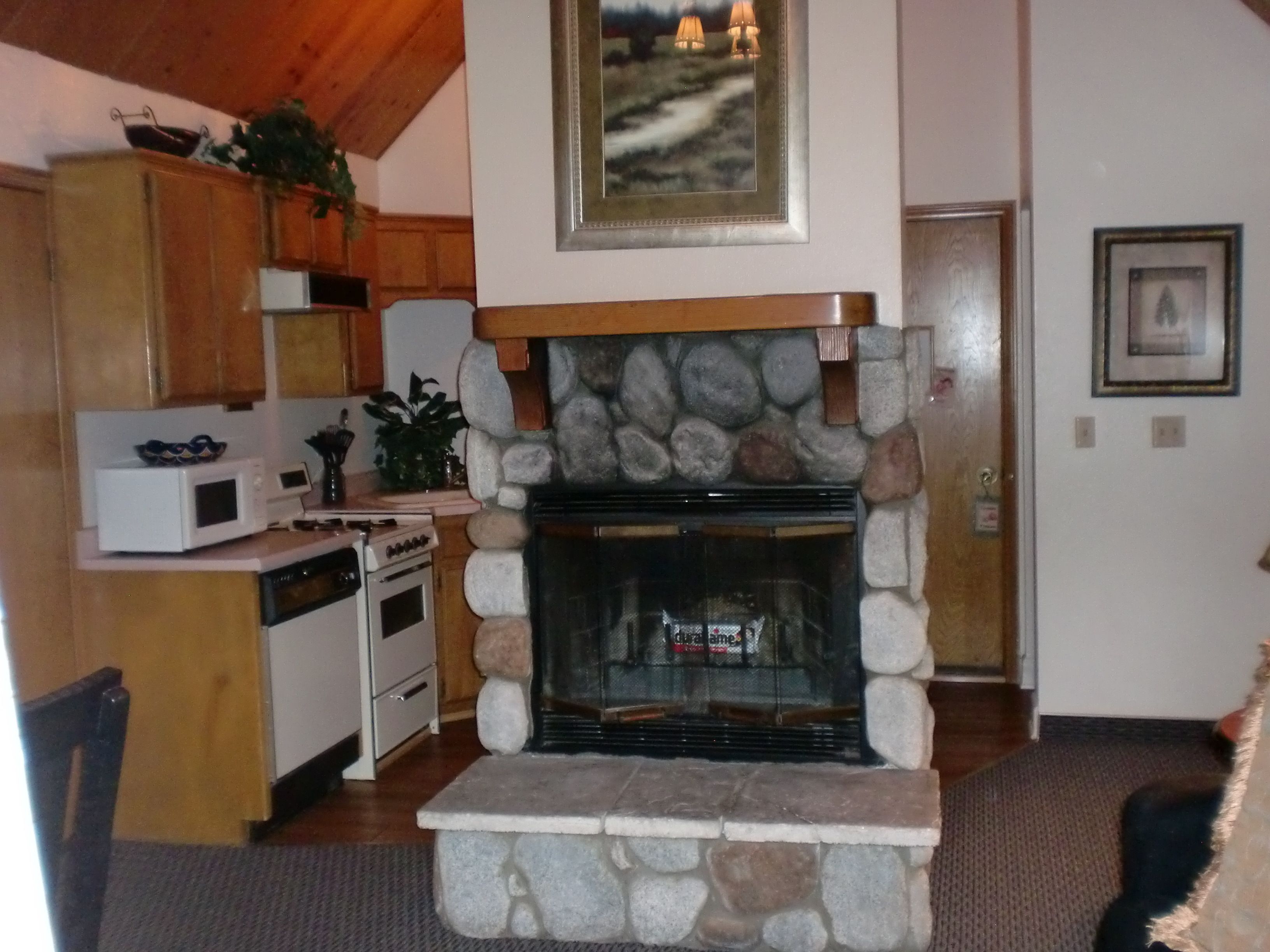 A stone fireplace with a wooden mantel, a kitchen with wooden cabinets and appliances, and a framed picture on the wall.