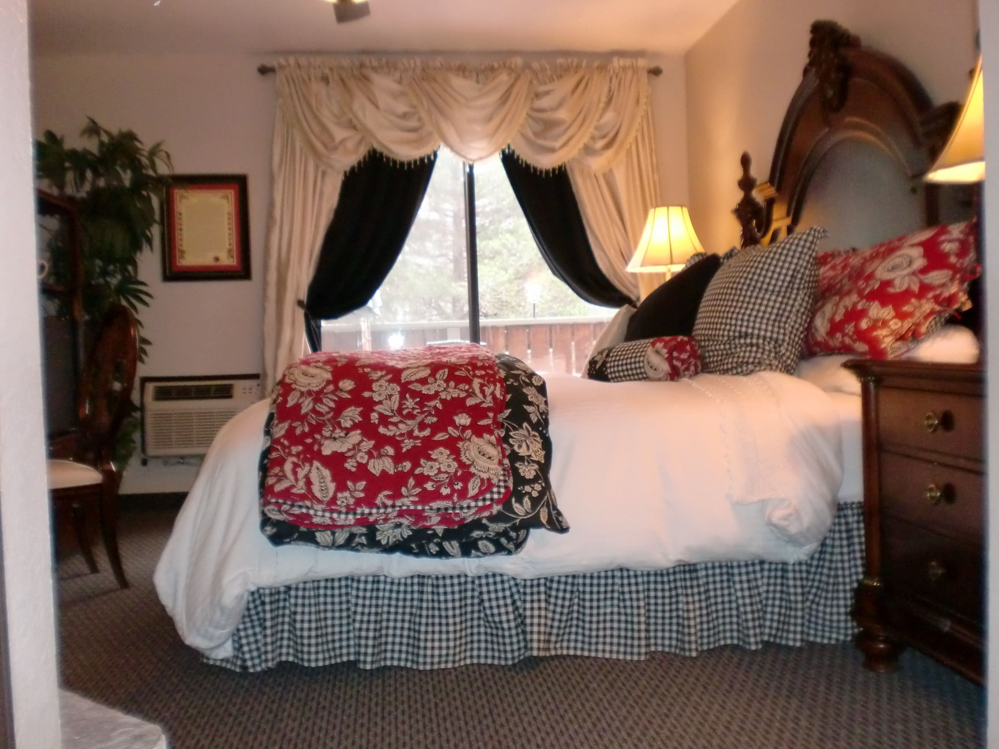 Un dormitorio con una cama grande, falda de cama a cuadros, colcha con motivos florales y varias almohadas. Se ve una ventana con cortinas, una mesita de noche y una lámpara.