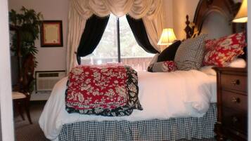Un dormitorio con una cama grande, falda de cama a cuadros, colcha con motivos florales y varias almohadas. Se ve una ventana con cortinas, una mesita de noche y una lámpara.
