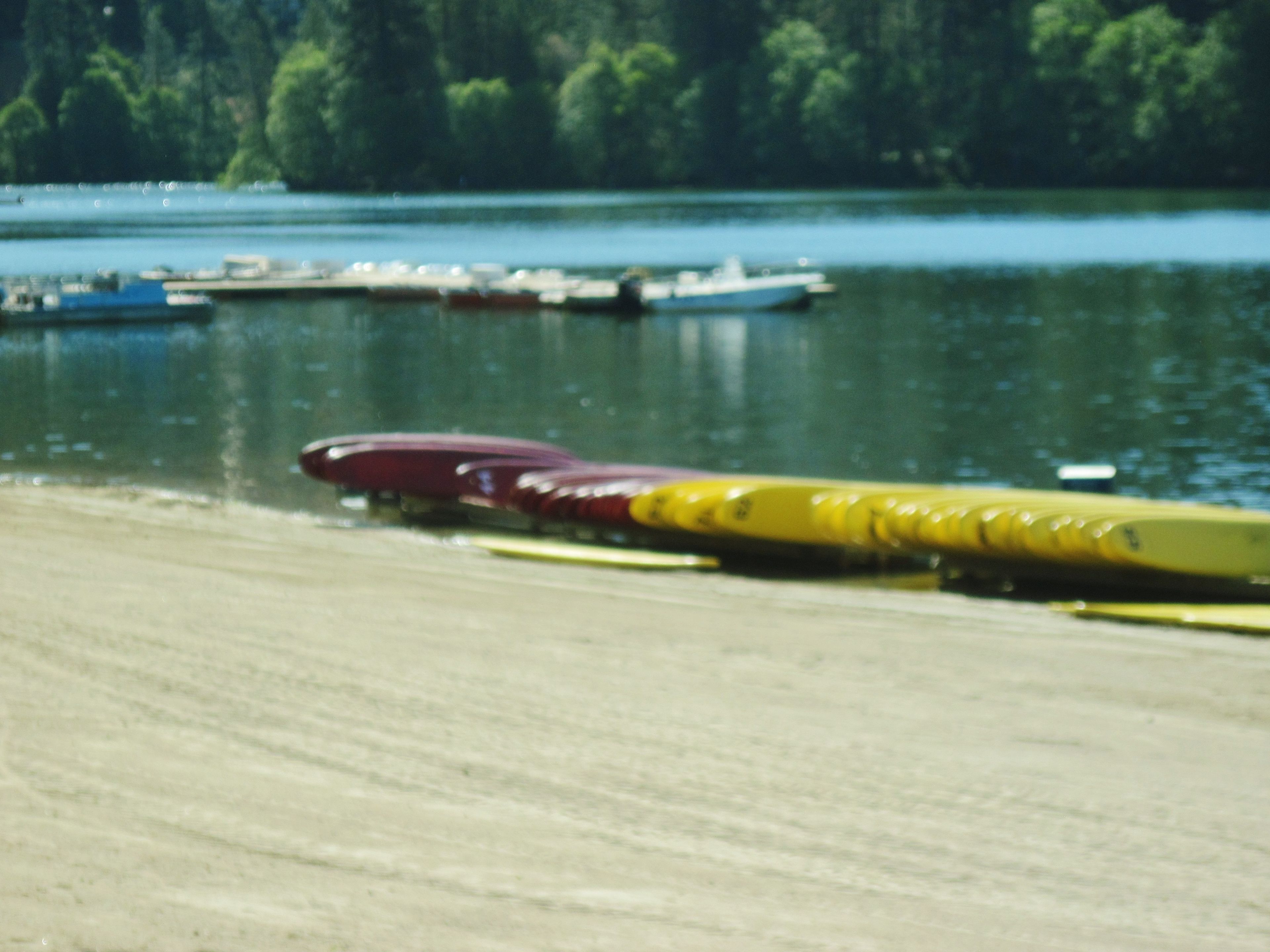 A row of canoes on a sandy shore by a calm lake with boats in the background.