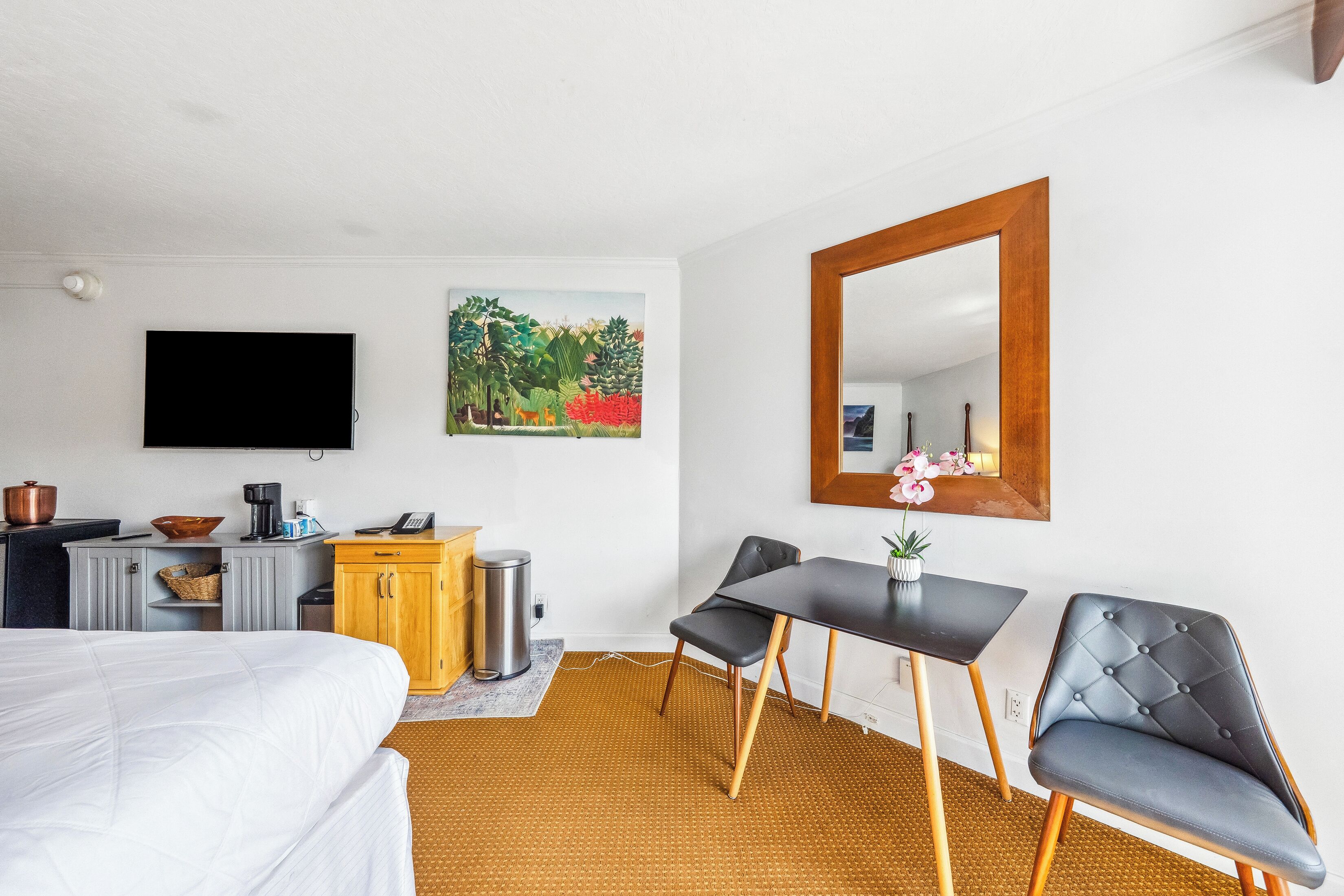 Una habitación de hotel con cama, escritorio, silla, televisor, cuadro y espejo.