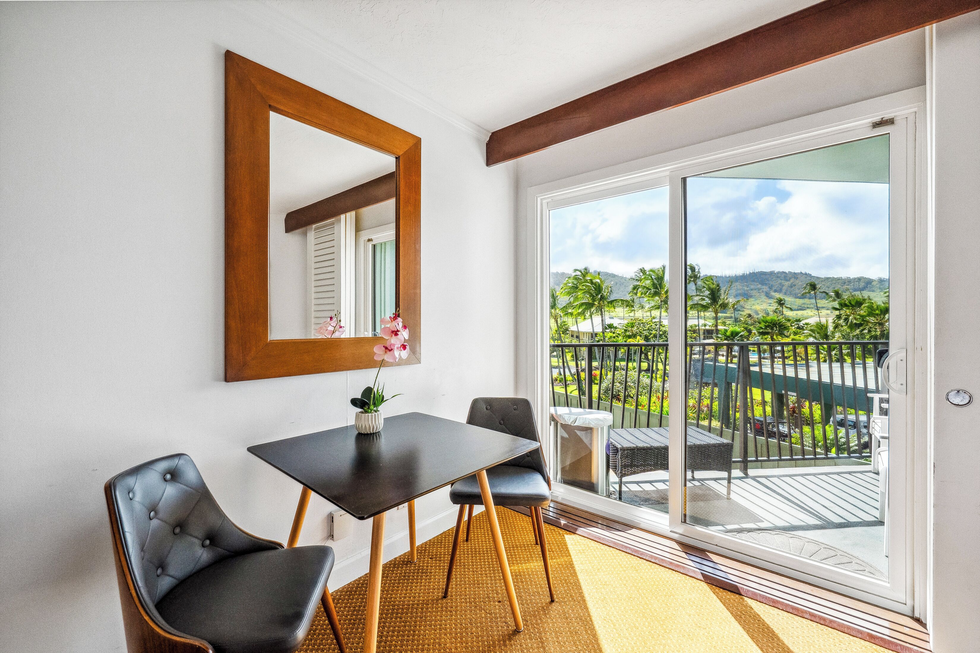 Un balcon avec vue sur les palmiers et les collines, une table avec des chaises et un grand miroir au mur.