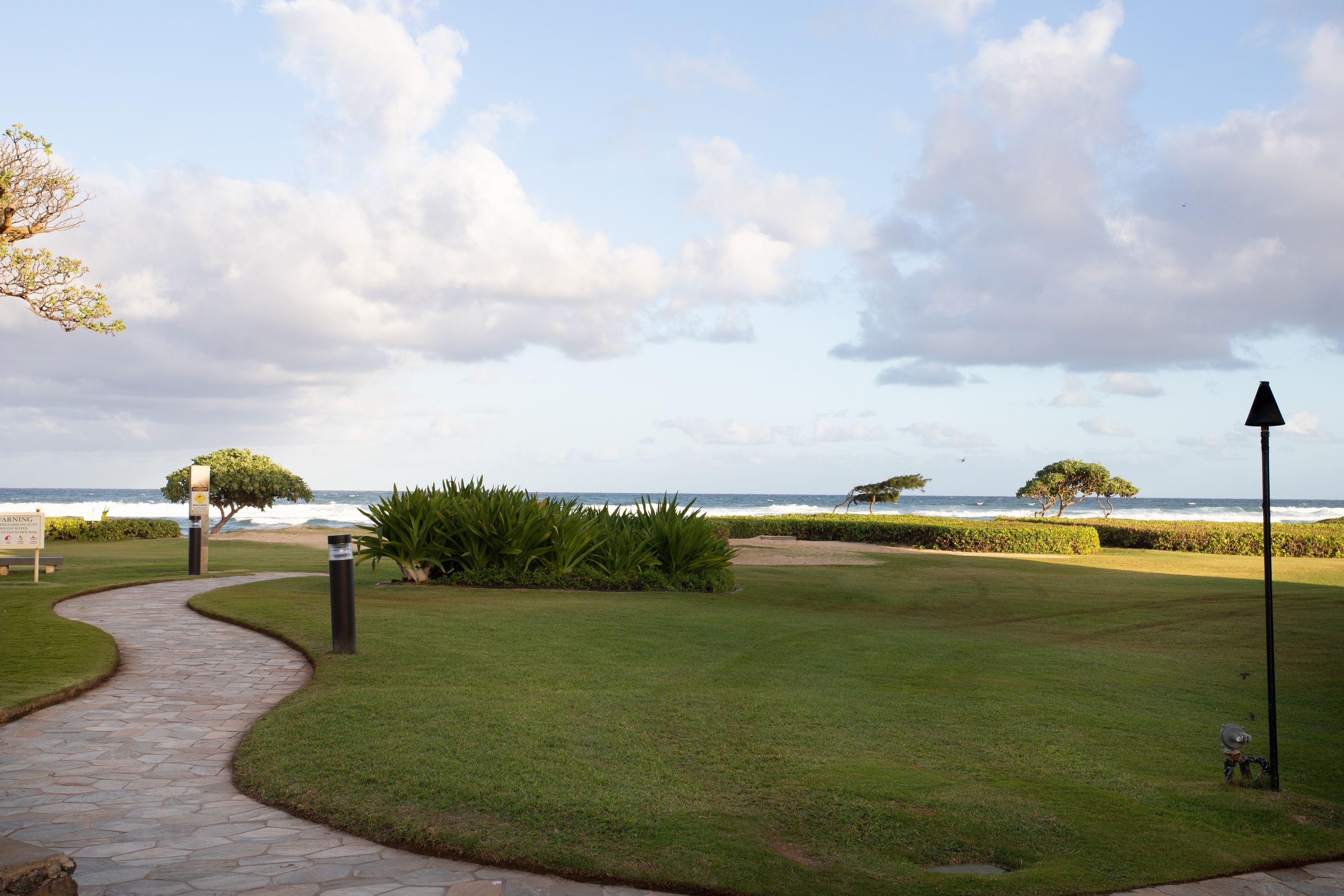 A pathway leading to a beach with a clear view of the ocean, a well-maintained lawn, and a few trees.
