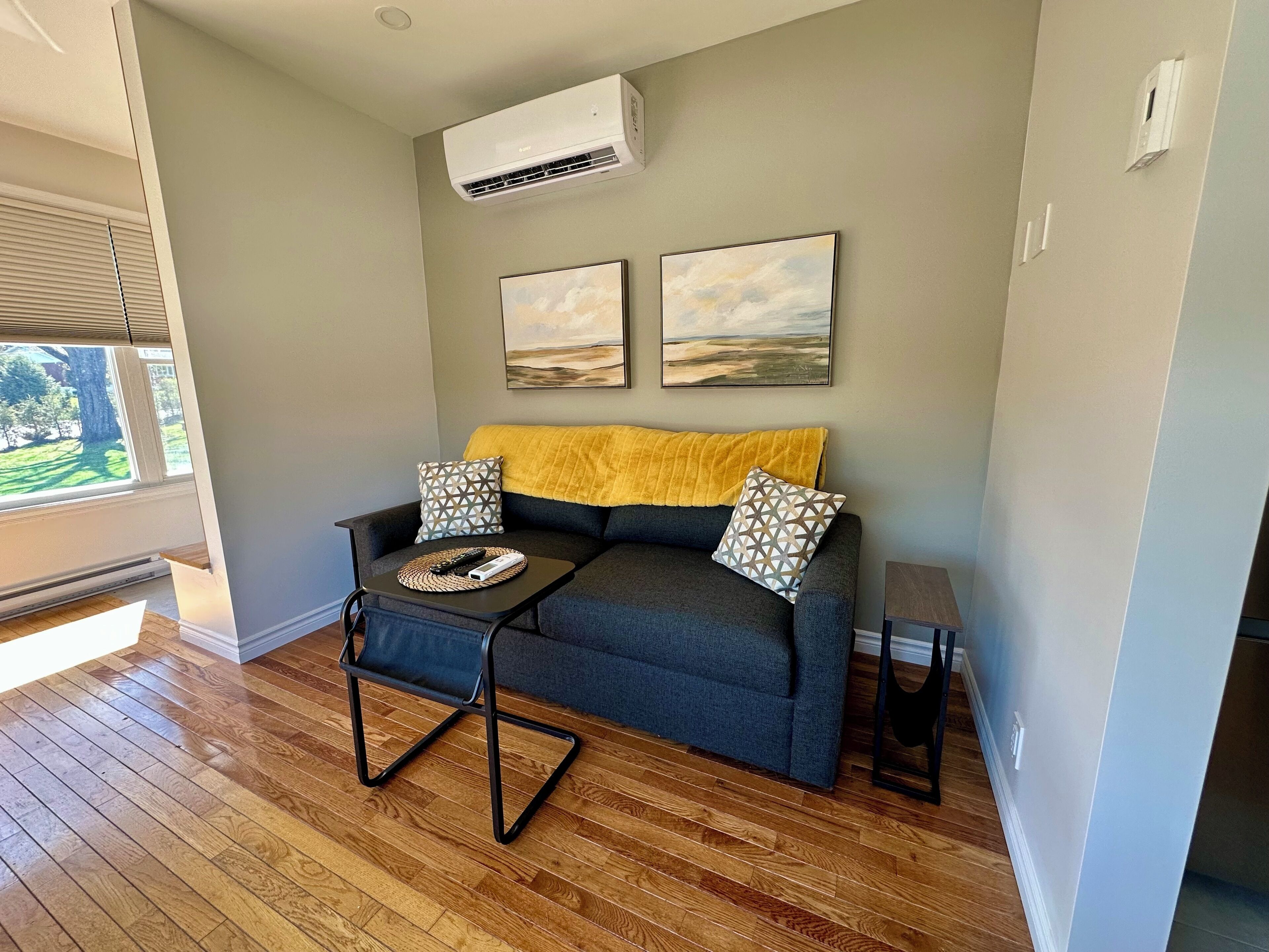 A living room with a grey sofa, two patterned pillows, a yellow throw, and a coffee table with remote controls.
