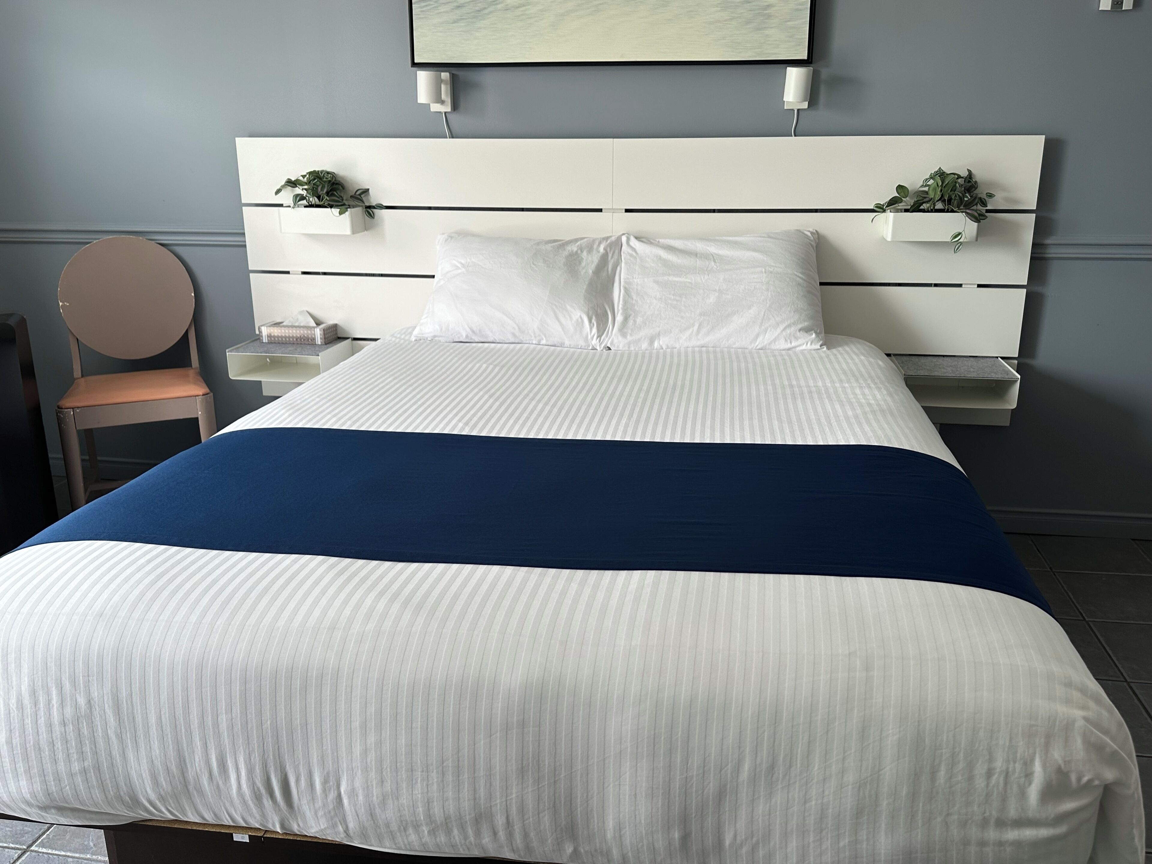 A neatly made bed with white and blue bedding, a white headboard with plant holders, and a small bedside table with a lamp.