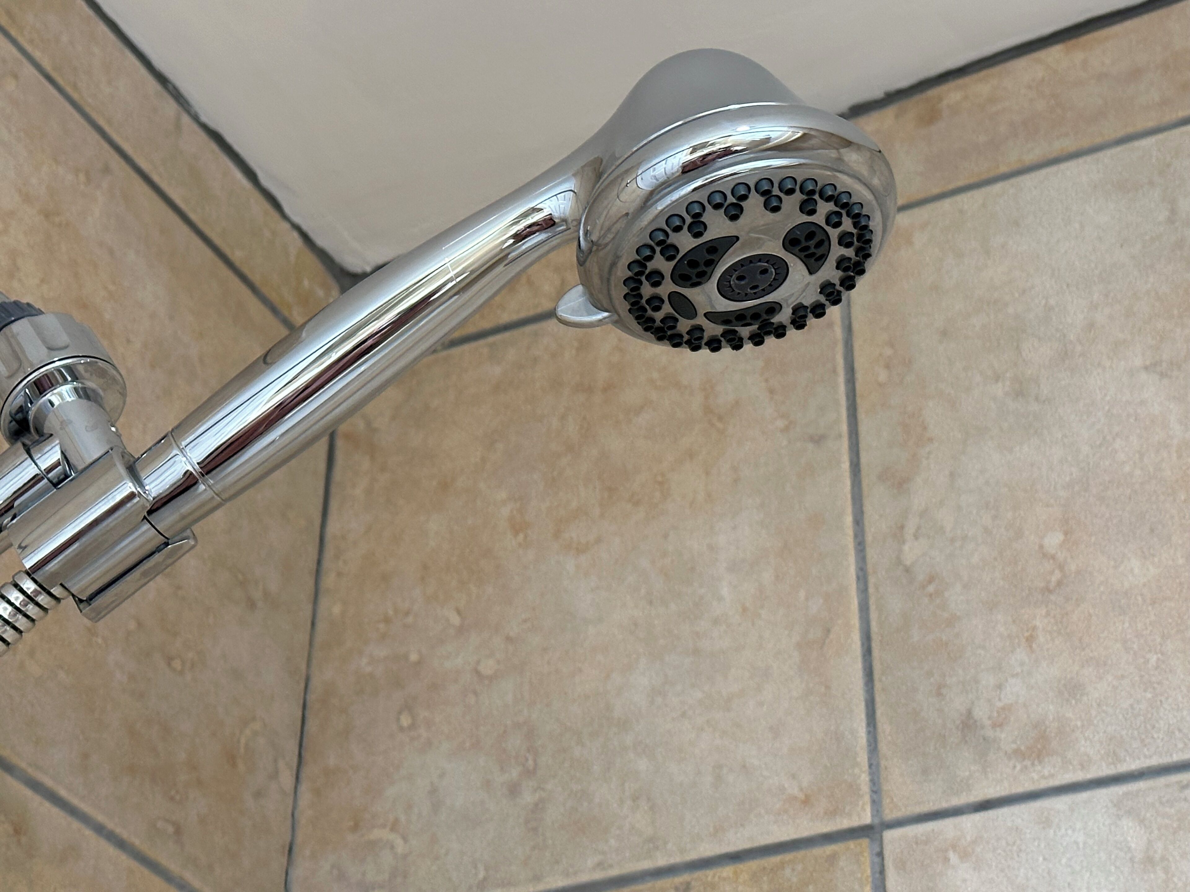 Um chuveiro cromado com padrão de spray circular, fixado numa parede com azulejos bege.