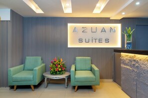 Un espace d’accueil avec une enseigne indiquant « AZUAN SUITES », ainsi que deux fauteuils verts, une petite table et un arrangement floral placé entre les deux.