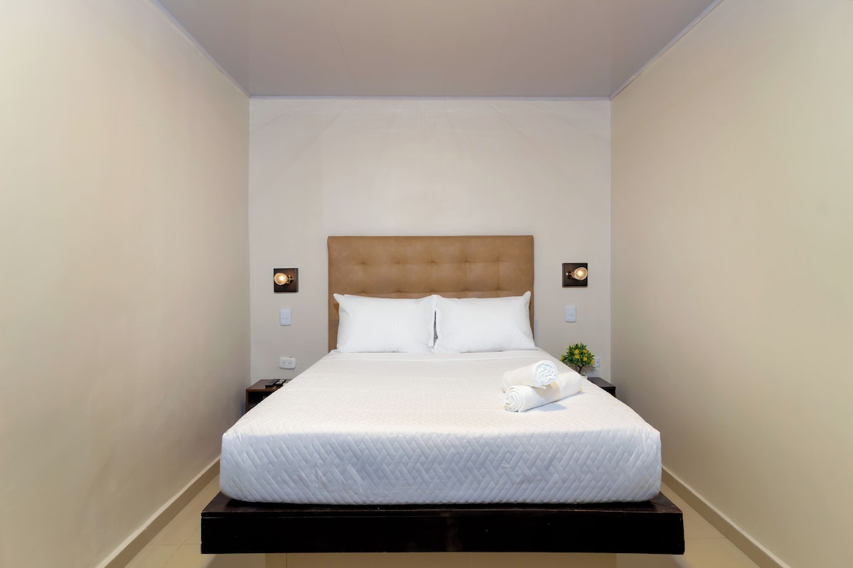 A hotel room with a bed, nightstands, a wall-mounted light, and a small plant.