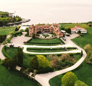 Una grande dimora, simile a un castello, con un prato curato, un viale a senso unico e una distesa d'acqua sullo sfondo.