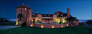 A large, castle-like building with a prominent tower, multiple chimneys, and a well-lit entrance. - OceanCliff Hotel (Newport)