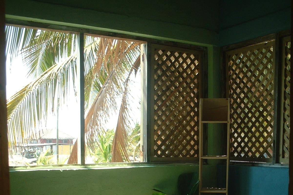 Un dormitorio con cama, una silla de madera, una mesita, una ventana con vista a palmeras y un estante de madera.