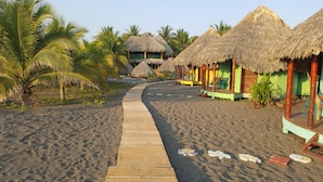 A wooden pathway leading to thatched-roof huts with green walls, surrounded by palm trees and sandy beach.