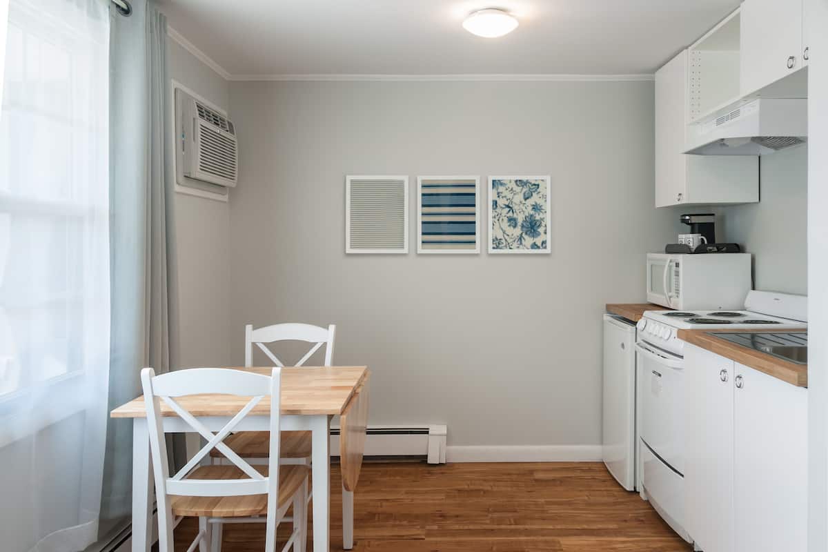 A small kitchen with white cabinets, a wooden table, and chairs. There are three framed pictures on the wall.