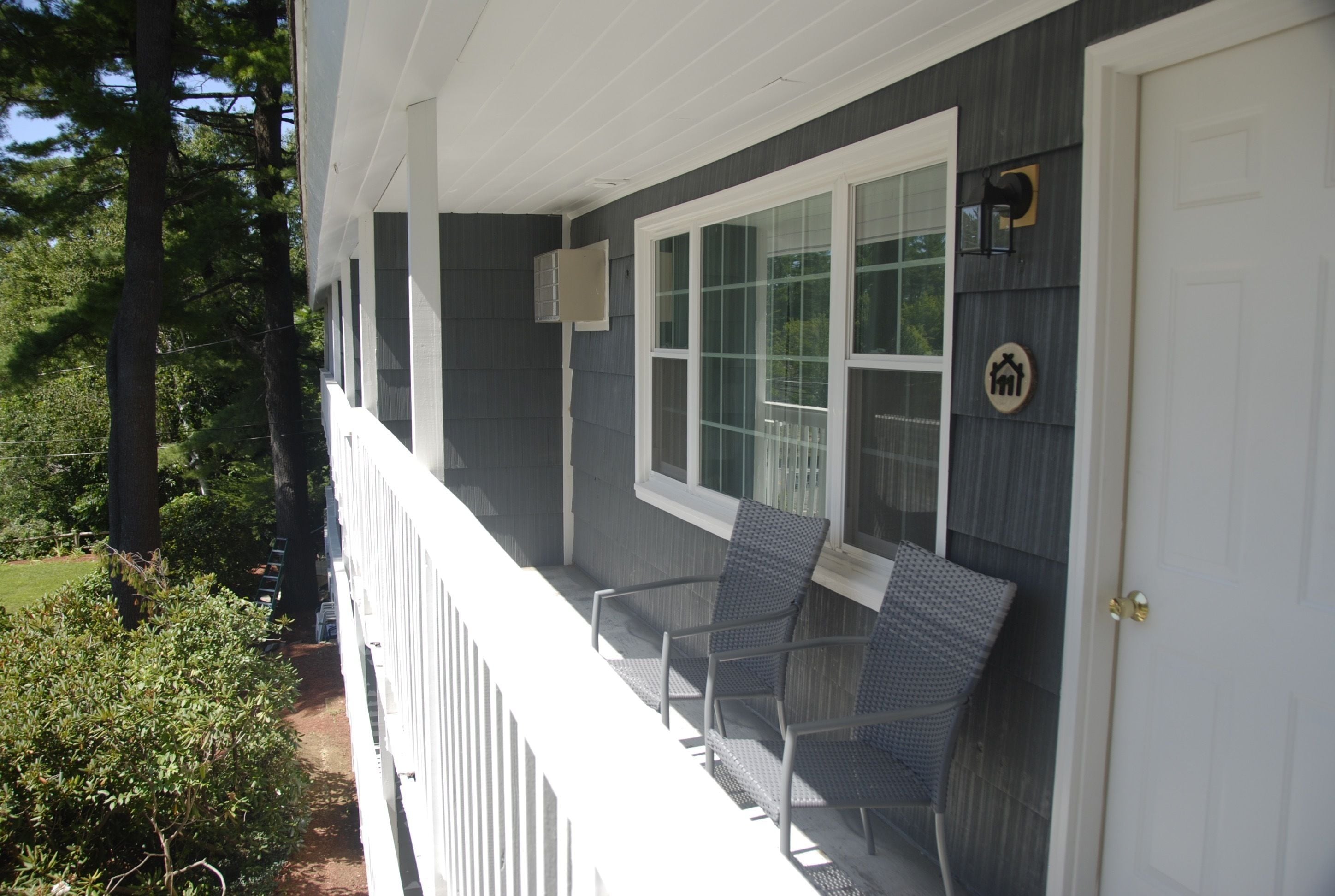 A porch with two wicker chairs, a white door, and a window with a view of trees.