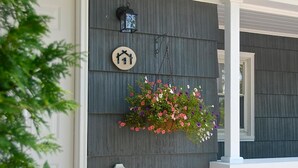 A house with a gray siding exterior, a white porch, a hanging flower basket, and a house number sign.