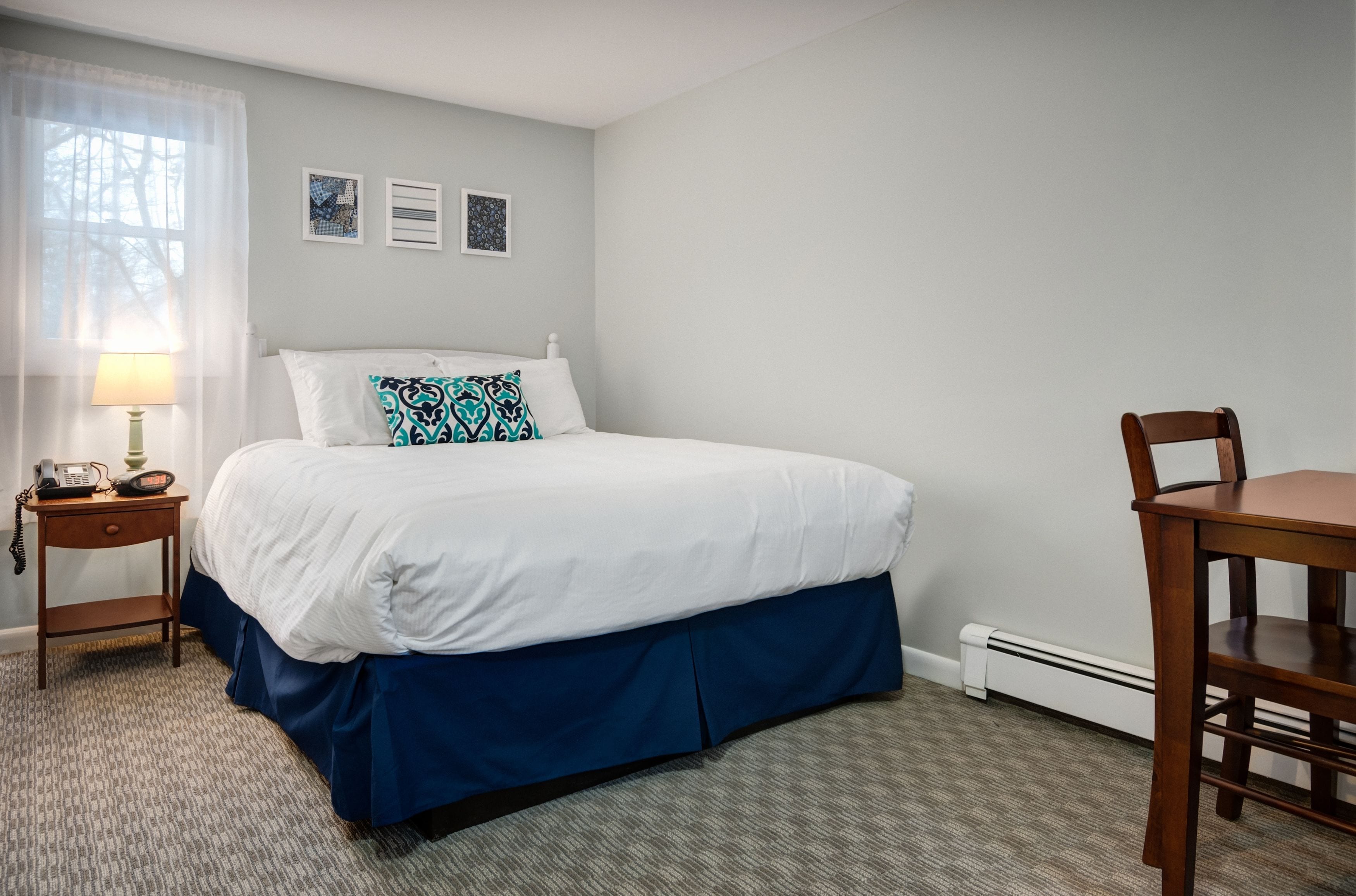 Habitación de hotel con una cama, una mesita de noche con lámpara, una silla y un cuadro con dos imágenes enmarcadas.