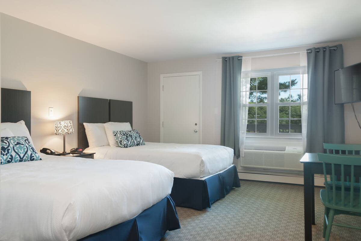 A hotel room with two beds, a desk, a chair, a TV, a window with curtains, and a door.