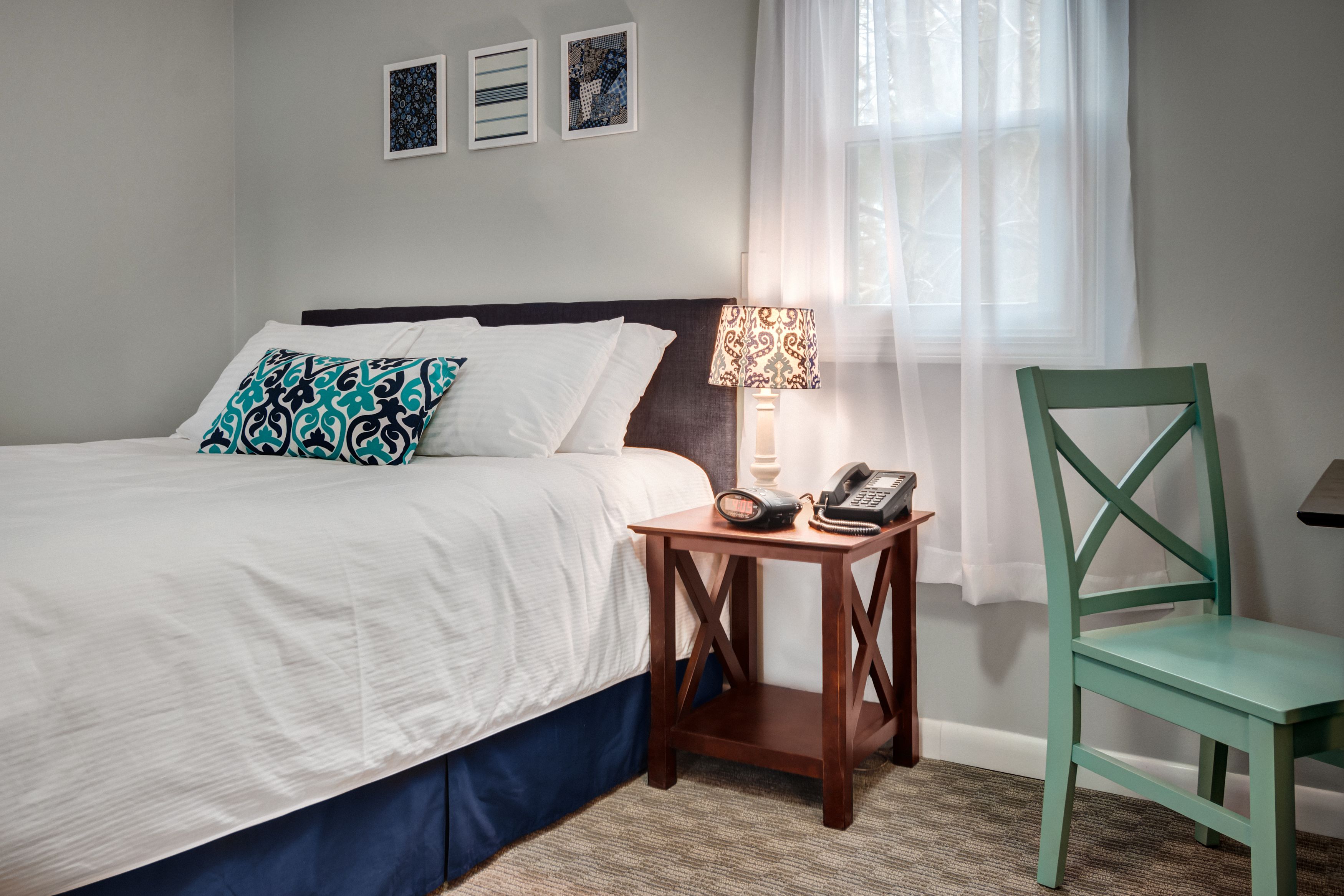 A neatly made bed with white bedding and patterned pillows, a bedside table with a lamp and phone, a green chair, and a window with sheer curtains.