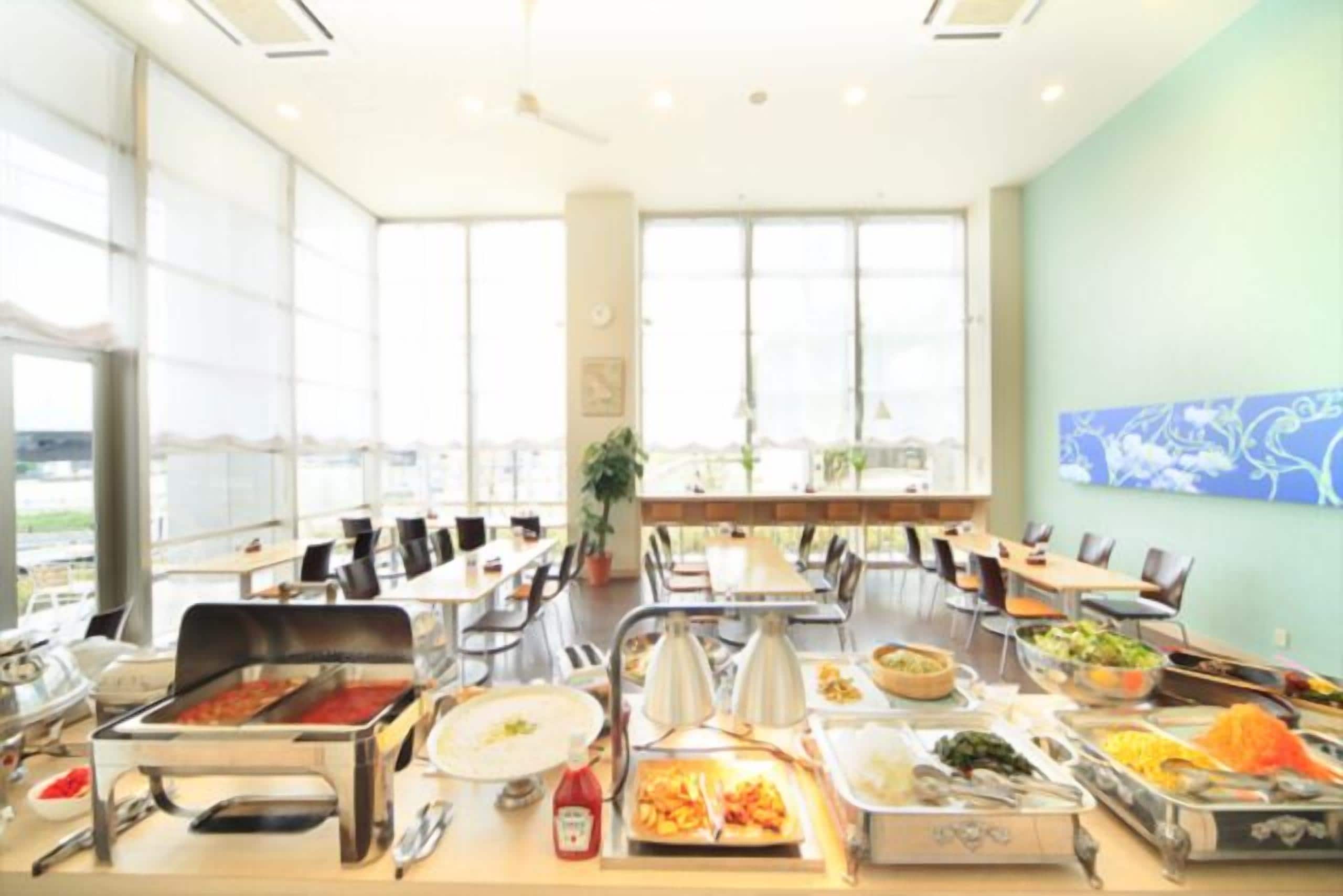 A spacious dining area with multiple tables and chairs, a buffet with various food items, and large windows providing natural light.