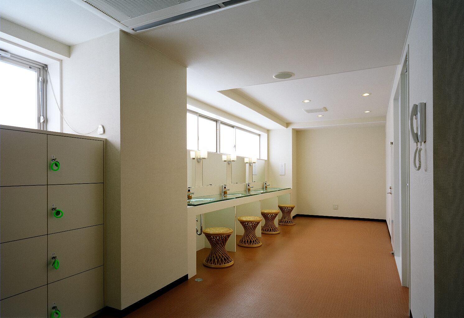 A hotel bathroom with a glass partition, multiple sinks, wicker stools, and a storage cabinet with green handles.