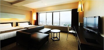 Habitación de hotel con cama, dos sofás, una mesita, televisor y ventana grande con vista a la ciudad.