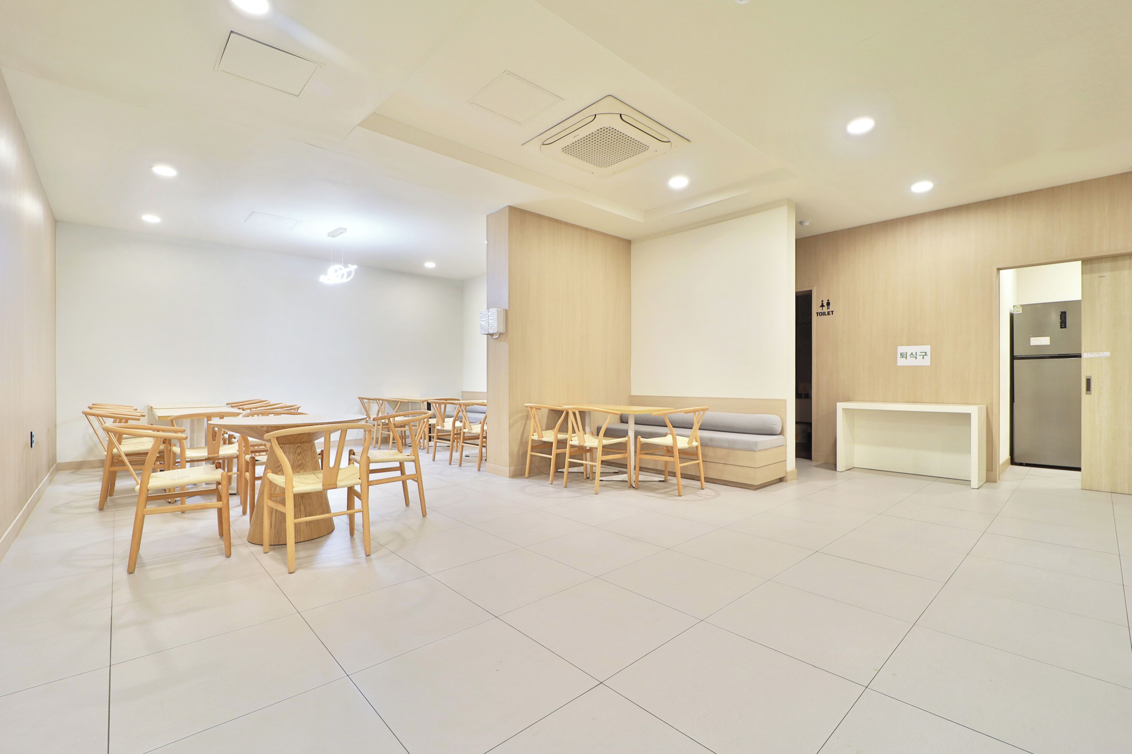 A spacious room with multiple wooden tables and chairs, a sofa, a refrigerator, and a toilet sign.
